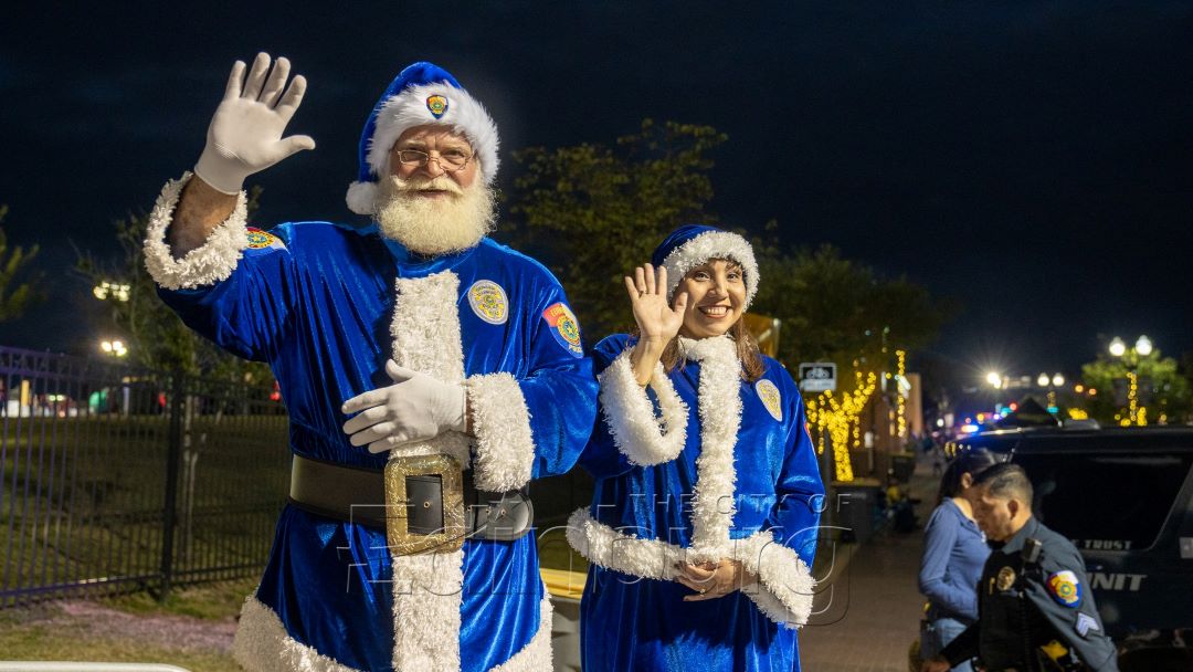 Two individuals dressed in blue Santa outfits standing outdoors at night, smiling and waving, with holiday lights and decorations visible in the background.