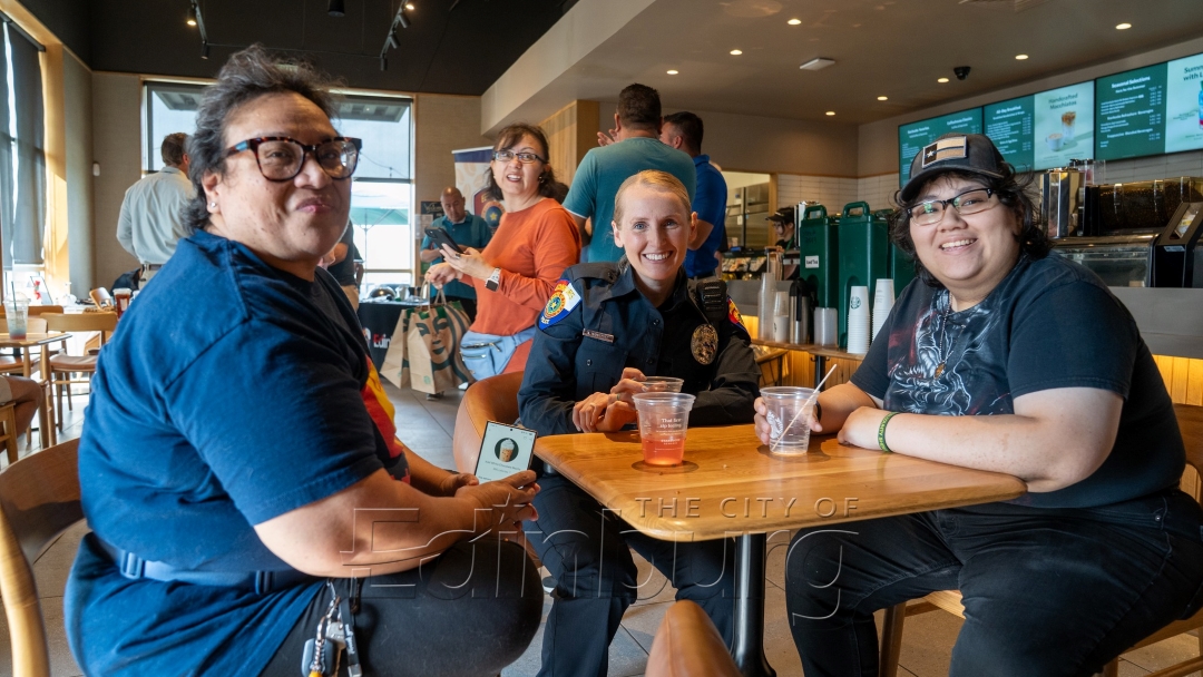 Police officer seated at a table inside a coffee shop with two community members, all smiling, with drinks and café seating visible in the background.