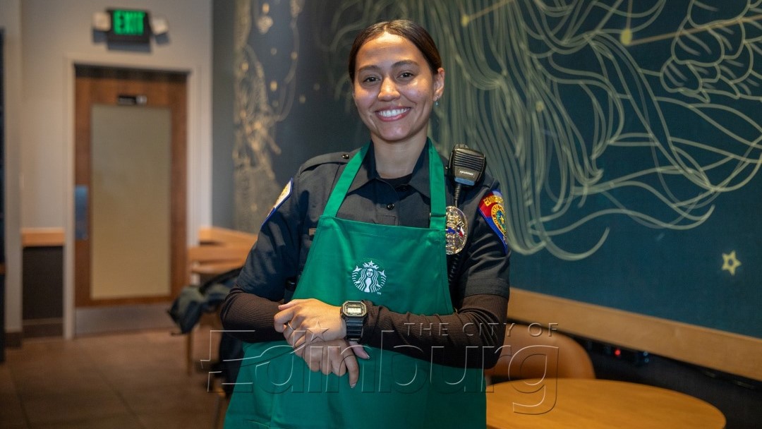 Police officer standing indoors wearing a green apron, smiling with arms crossed inside a coffee shop setting.