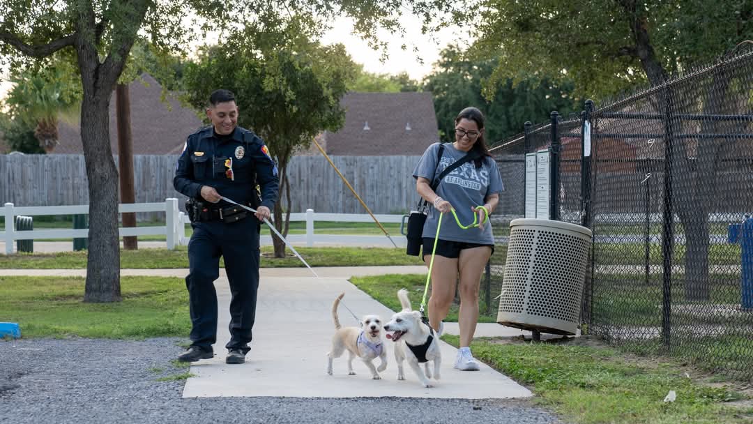 Police officer walking alongside a community member who is holding two small dogs on leashes along a sidewalk in a park.