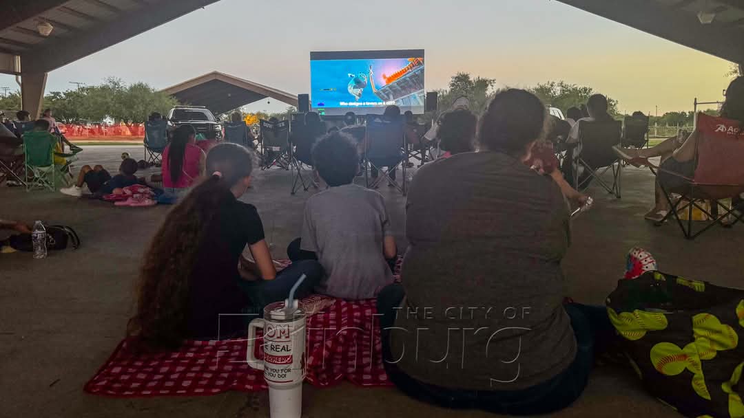 Group of people sitting on blankets under a covered area watching a movie projected on a large outdoor screen.