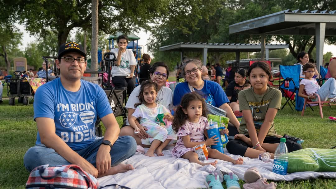 Family sitting on a blanket in a park with children and adults facing the camera, with other attendees and a pavilion visible in the background.