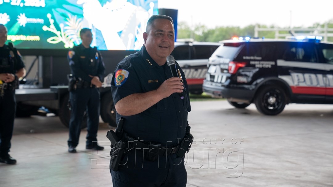 Police officer holding a microphone and speaking to a crowd at an outdoor event, with a patrol vehicle and other officers visible behind him.