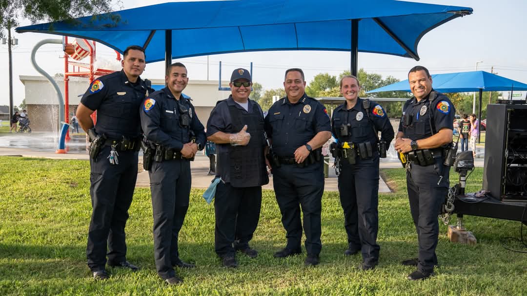 Group of uniformed police officers standing together outdoors on a grassy area under a blue shade structure, posing for a photo.