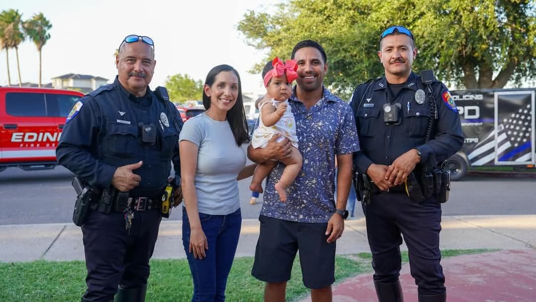 Two Edinburg police officers in uniform stand outdoors with a smiling man and woman holding a baby in a pink bow. A red Edinburg Fire Department truck and a police trailer are visible in the background.